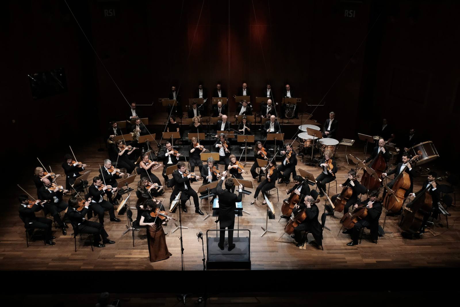 L'Orchestra della Svizzera italiana in concerto sul palco, vista dall'alto con il direttore e una violinista solista in primo piano: tra gli obiettivi del nuovo corso, il rafforzamento dell'identità interpretativa e una pianificazione pluriennale del repertorio.